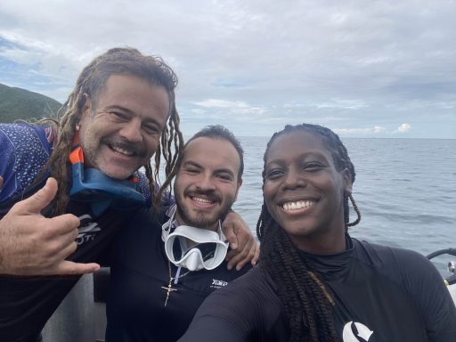 Three people smiling taking a 'selfie' photo. The first person on the far left is doing the "hang loose" sign with their right and and their
